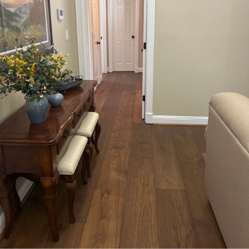 A wooden console table with two upholstered stools sits in a hallway with hardwood floors. A vase of yellow flowers and a decorative tray are on the table.